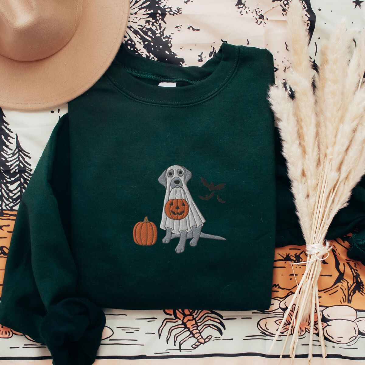 Silver Lab Ghost Sweatshirt - Forest Green Forest Green crewneck sweatshirt laid flat with a felt hat and dried pampas grass, embroidered with a silver Labrador under a white ghost sheet beside a pumpkin and bats