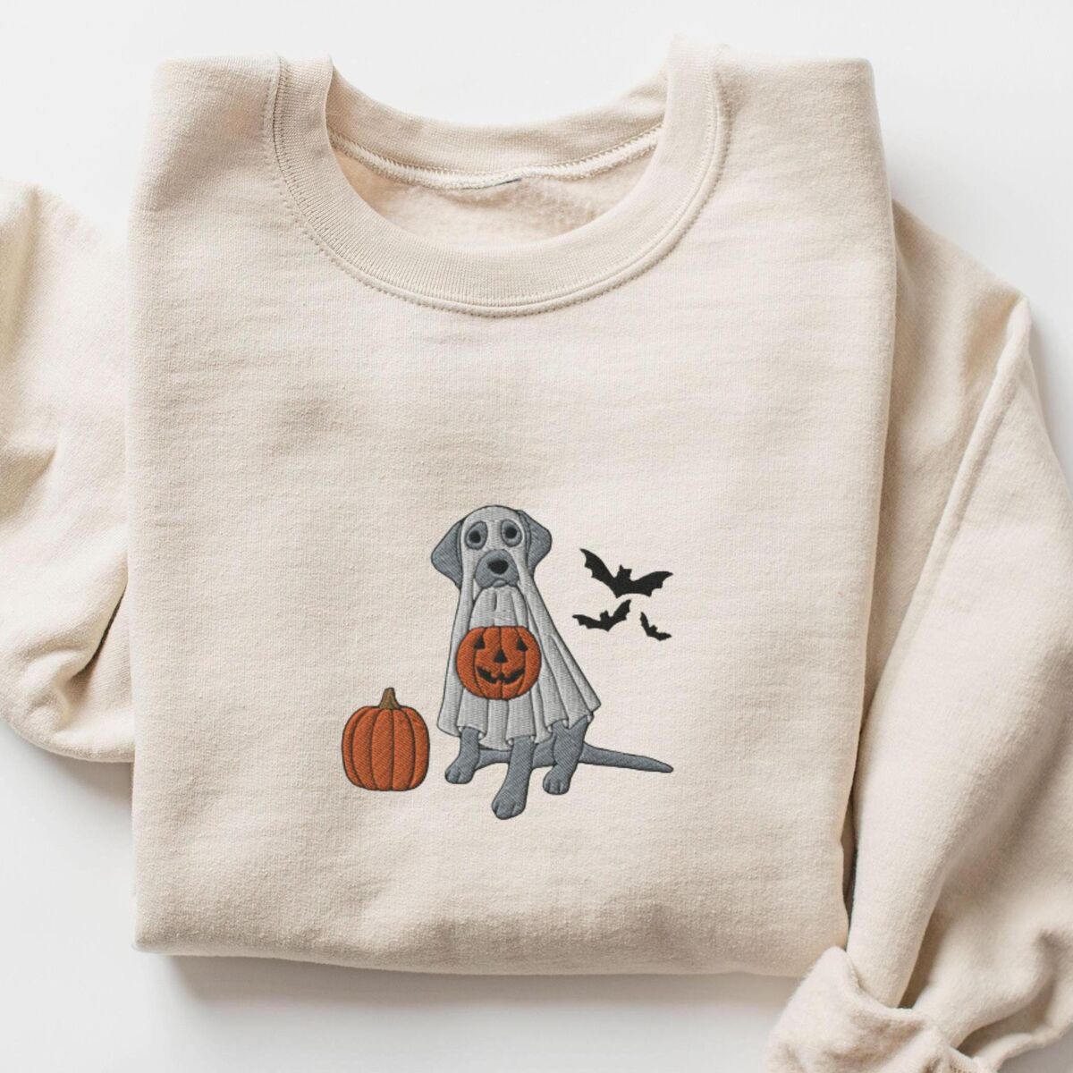 Silver Lab Ghost Sweatshirt - Sand Sand crewneck sweatshirt laid flat on a white background, embroidered with a silver Labrador in a white ghost sheet holding a jack-o'-lantern bucket beside a pumpkin and three bats