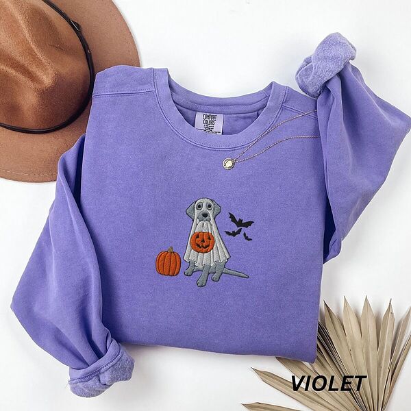 Violet crewneck sweatshirt laid flat with a brown felt hat and a dried palm leaf, embroidered with a silver Labrador in a white ghost sheet holding a jack-o'-lantern bucket beside a pumpkin and three bats