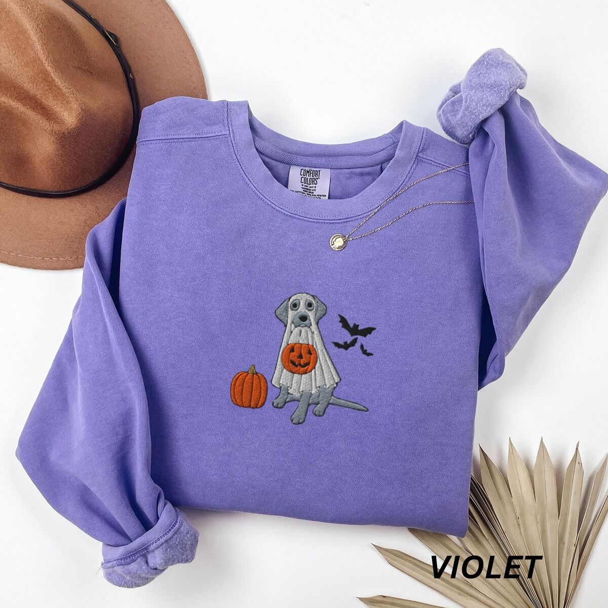 Silver Lab Ghost Sweatshirt – Violet Violet crewneck sweatshirt laid flat with a brown felt hat and a dried palm leaf, embroidered with a silver Labrador in a white ghost sheet holding a jack-o'-lantern bucket beside a pumpkin and three bats