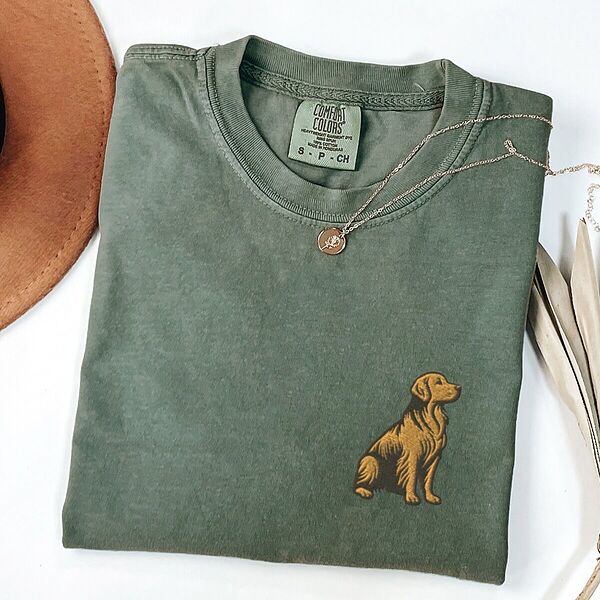 Moss T-shirt laid flat with a necklace and a brown hat, embroidered with a small sitting golden retriever on the left chest