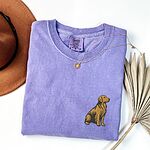 Violet T-shirt laid flat with a necklace and dried palm leaves, embroidered with a small sitting golden retriever on the left chest