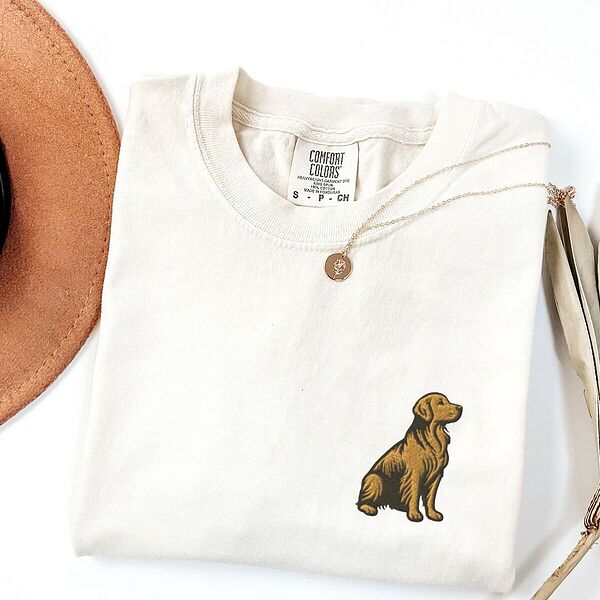 White T-shirt laid flat with a necklace and a brown hat, embroidered with a small sitting golden retriever on the left chest