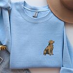 Light Blue crewneck sweatshirt laid flat beside a tan hat and jeans, embroidered with a small sitting golden retriever on the left chest