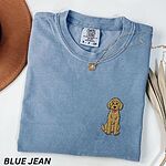 Blue Jean T-shirt laid flat with a hat and a necklace, embroidered with a small golden goldendoodle sitting on the left chest