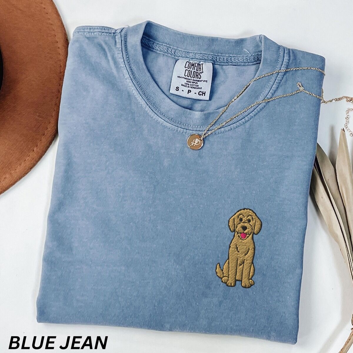Sitting Goldendoodle Shirt – Blue Jean Blue Jean T-shirt laid flat with a hat and a necklace, embroidered with a small golden goldendoodle sitting on the left chest