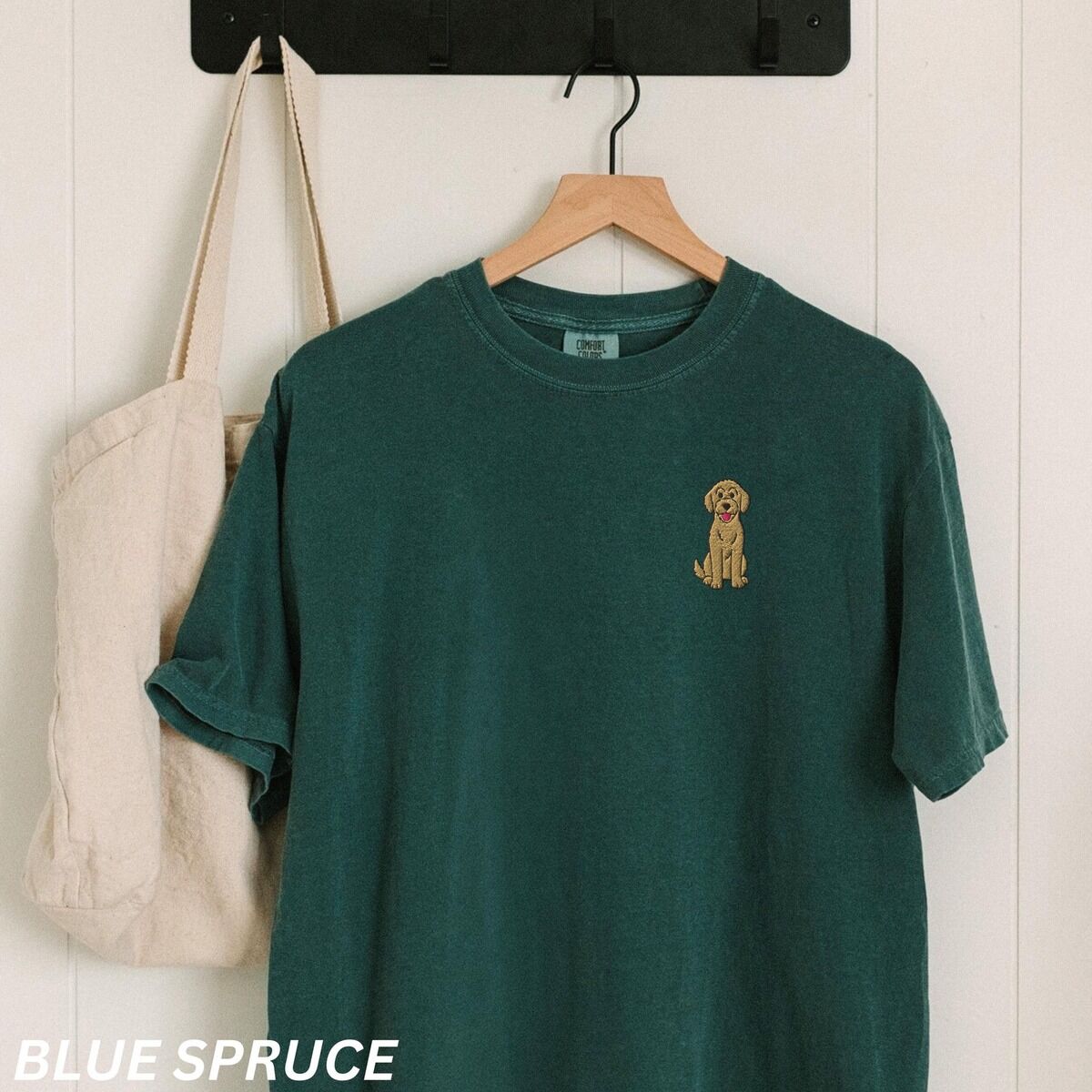 Sitting Goldendoodle Shirt – Blue Spruce Blue Spruce T-shirt on a hanger beside a canvas tote, embroidered with a small golden goldendoodle sitting on the left chest