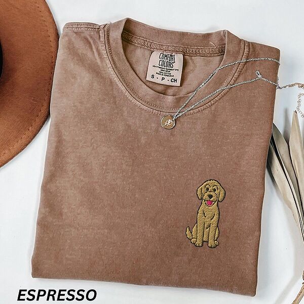 Espresso T-shirt laid flat with a hat and a necklace, embroidered with a small golden goldendoodle sitting on the left chest