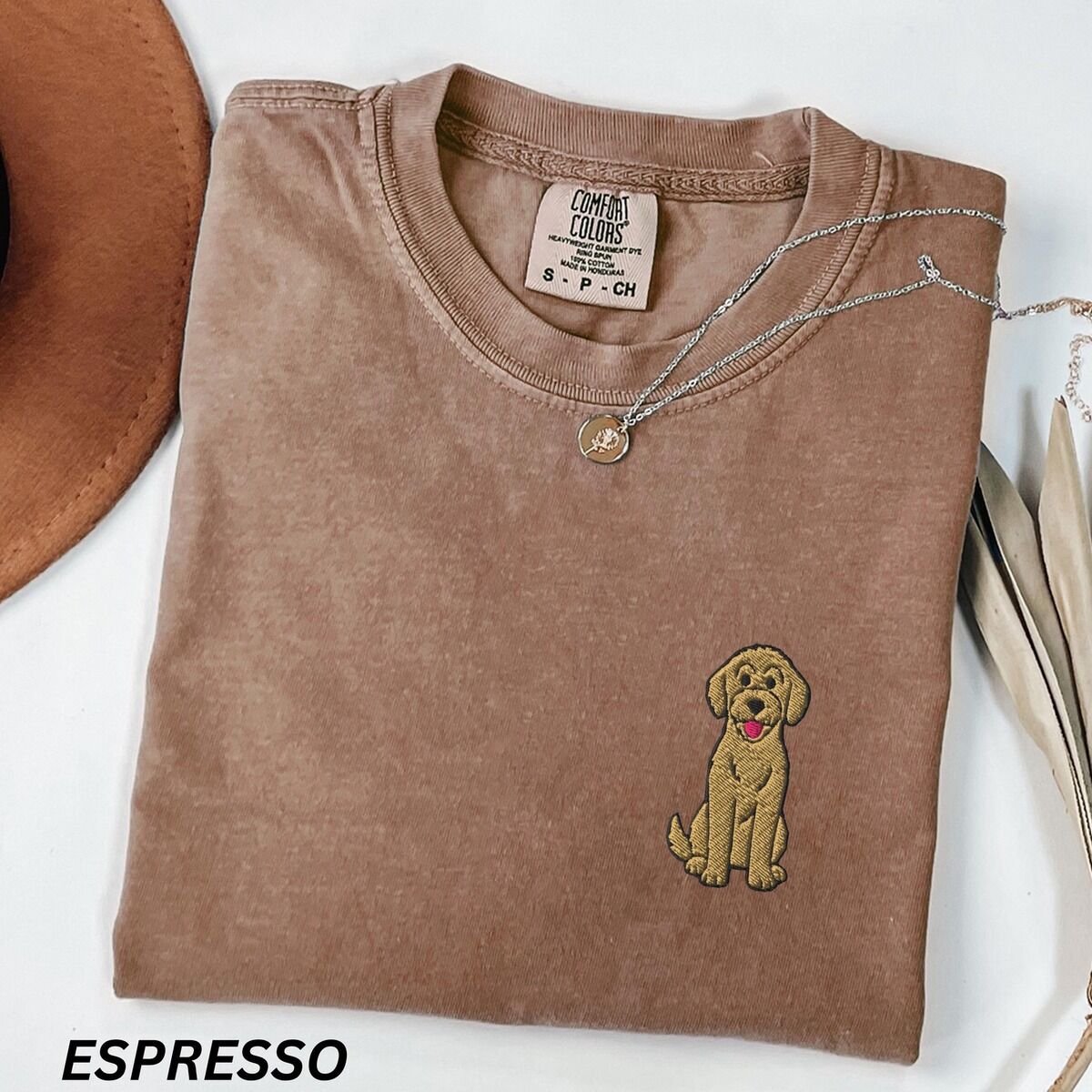 Sitting Goldendoodle Shirt – Espresso Espresso T-shirt laid flat with a hat and a necklace, embroidered with a small golden goldendoodle sitting on the left chest