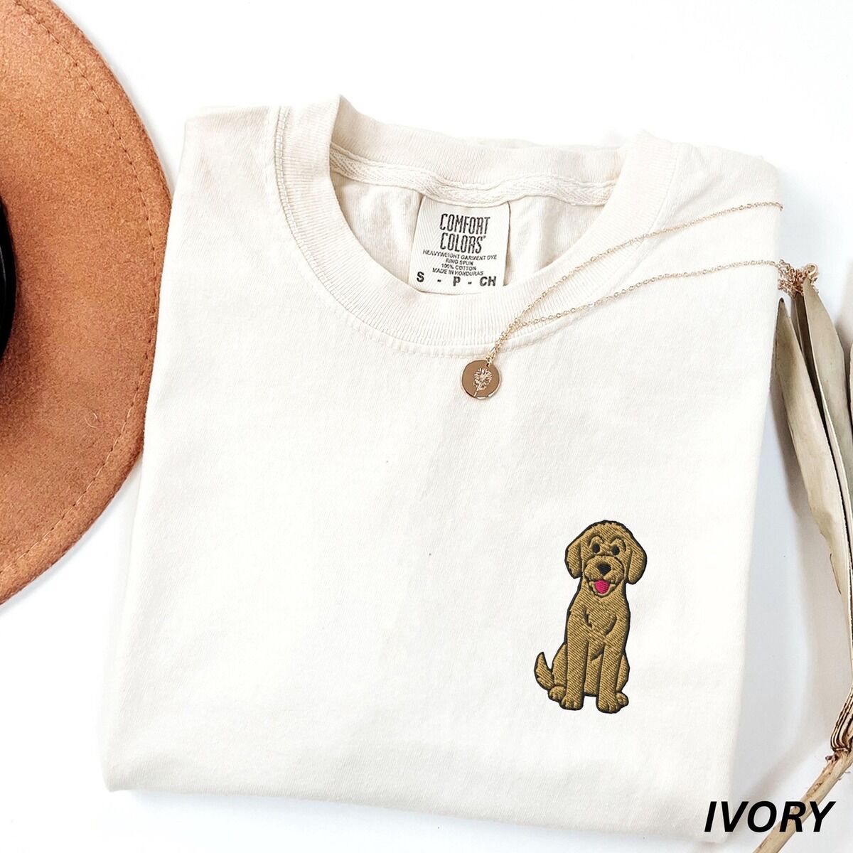 Sitting Goldendoodle Shirt – Ivory Ivory T-shirt laid flat with a hat and a necklace, embroidered with a small golden goldendoodle sitting on the left chest