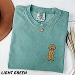 Light Green T-shirt laid flat with a hat and a necklace, embroidered with a small golden goldendoodle sitting on the left chest