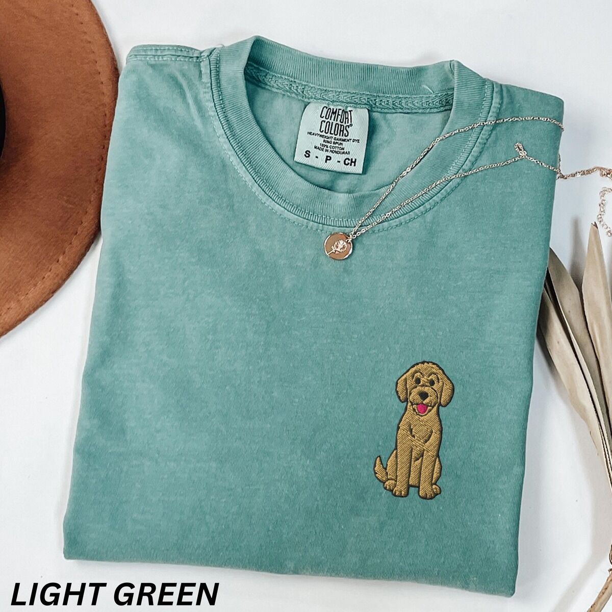Sitting Goldendoodle Shirt – Light Green Light Green T-shirt laid flat with a hat and a necklace, embroidered with a small golden goldendoodle sitting on the left chest