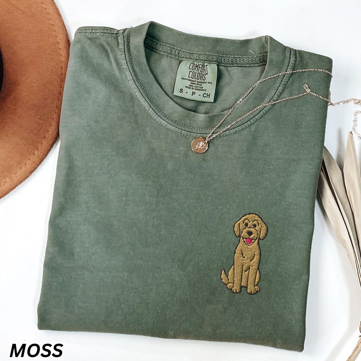 Sitting Goldendoodle Shirt – Moss Moss T-shirt laid flat with a hat and a necklace, embroidered with a small golden goldendoodle sitting on the left chest