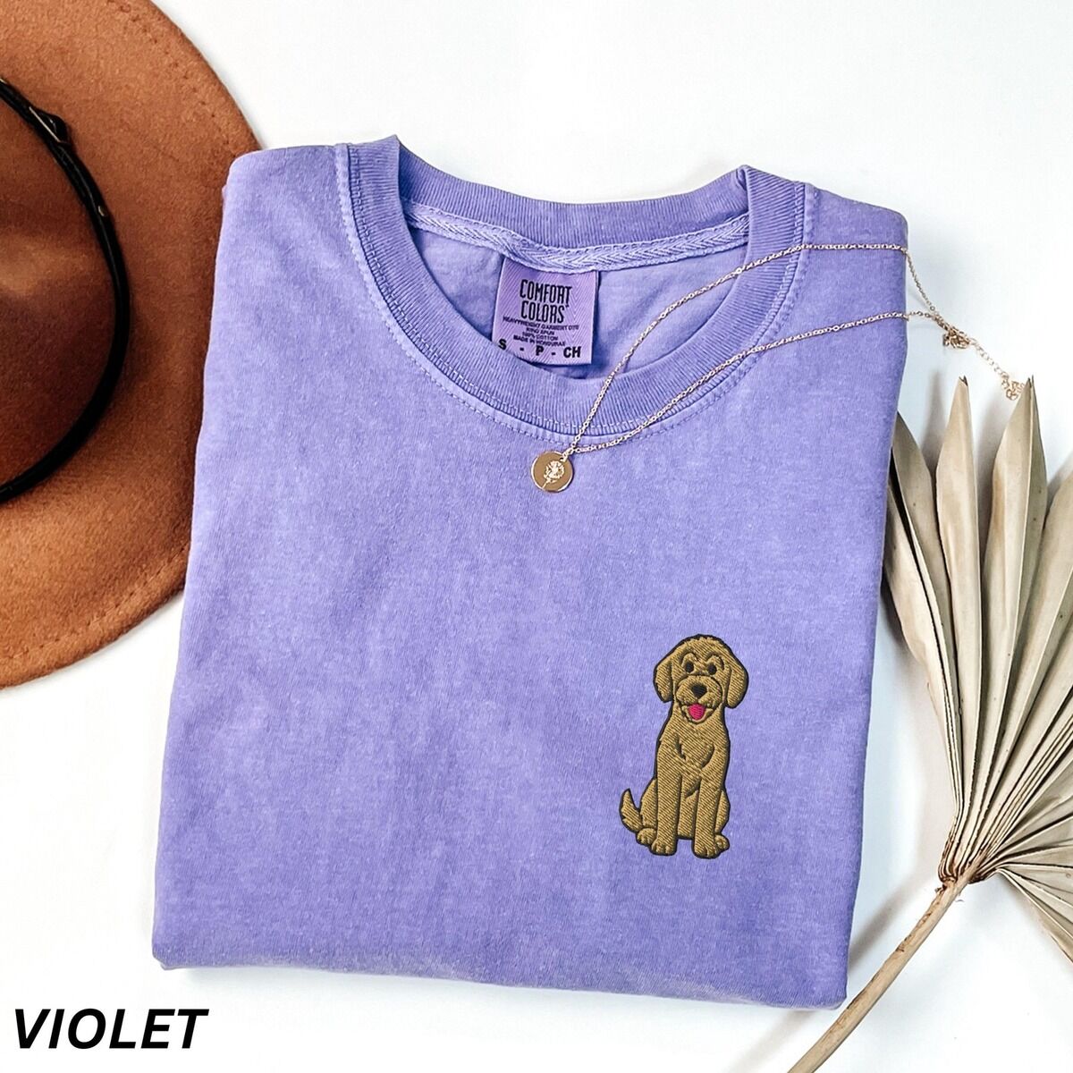 Sitting Goldendoodle Shirt – Violet Violet T-shirt laid flat with a hat and dried palm leaves, embroidered with a small golden goldendoodle sitting on the left chest
