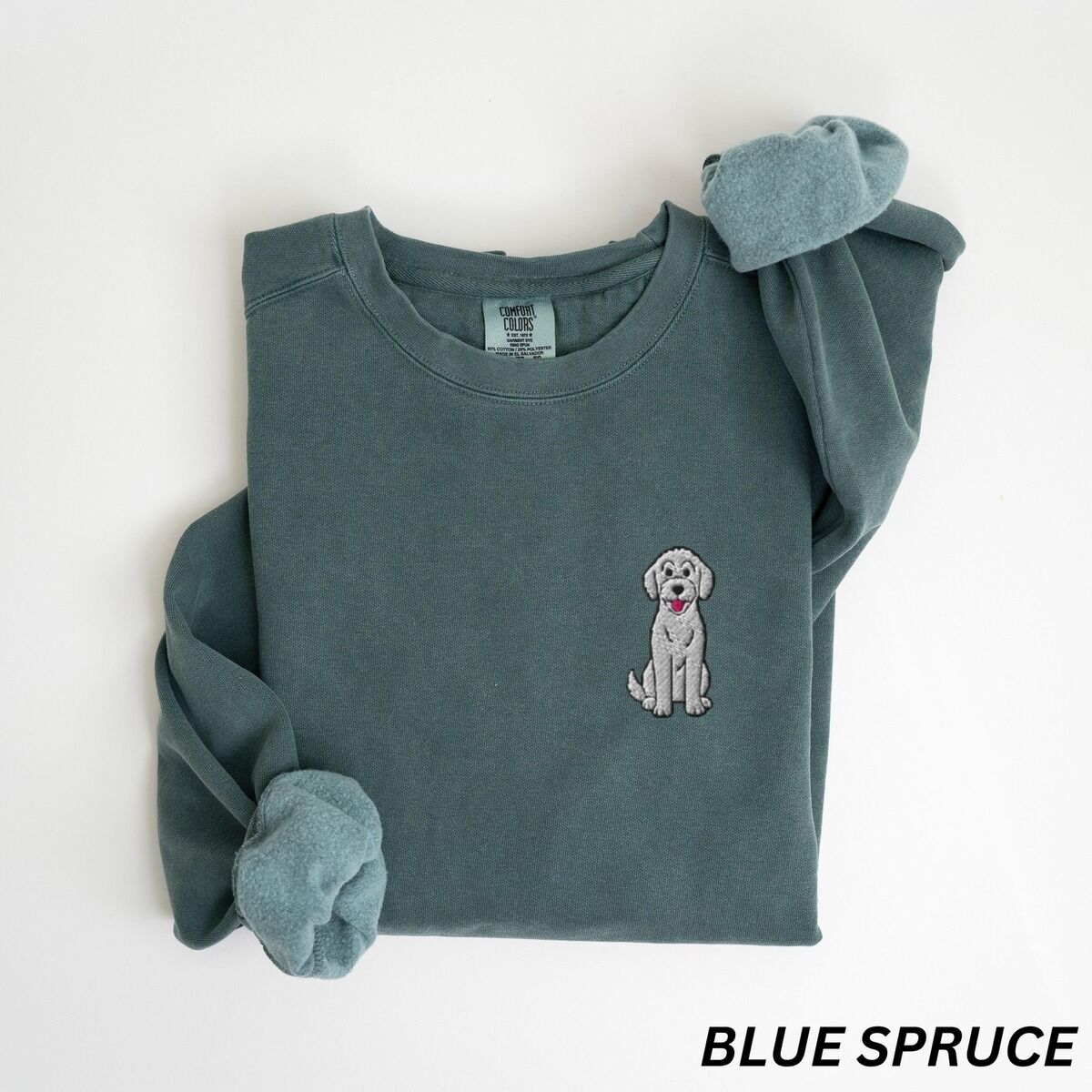 Sitting Goldendoodle Sweatshirt – Blue Spruce Blue Spruce crewneck sweatshirt laid flat, embroidered with a small light grey goldendoodle sitting on the left chest