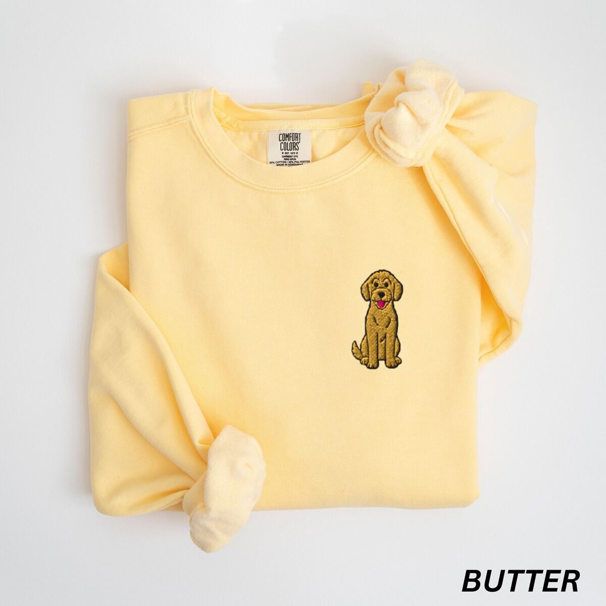 Sitting Goldendoodle Sweatshirt – Butter Butter crewneck sweatshirt laid flat with a knotted sleeve, embroidered with a small golden goldendoodle sitting on the left chest