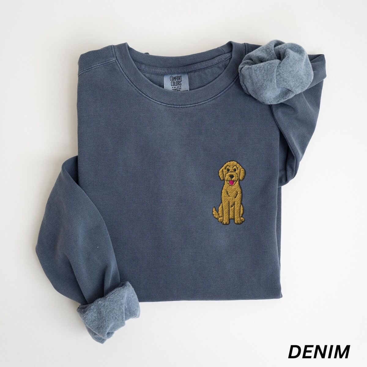 Sitting Goldendoodle Sweatshirt – Denim Denim crewneck sweatshirt laid flat with a rolled cuff, embroidered with a small golden goldendoodle sitting on the left chest