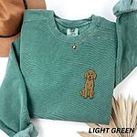 Light Green crewneck sweatshirt laid flat with a hat and a necklace, embroidered with a small golden goldendoodle sitting tall on the left chest