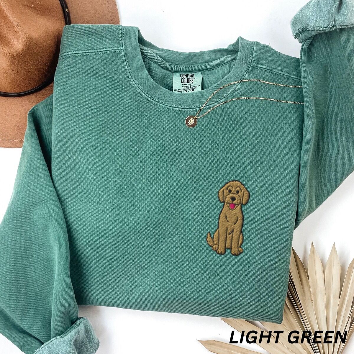 Sitting Goldendoodle Sweatshirt – Light Green Light Green crewneck sweatshirt laid flat with a hat and a necklace, embroidered with a small golden goldendoodle sitting tall on the left chest