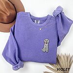 Violet crewneck sweatshirt laid flat with a hat and dried palm leaves, embroidered with a small light grey goldendoodle sitting on the left chest