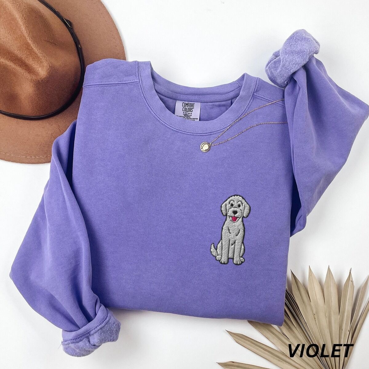 Sitting Goldendoodle Sweatshirt – Violet Violet crewneck sweatshirt laid flat with a hat and dried palm leaves, embroidered with a small light grey goldendoodle sitting on the left chest