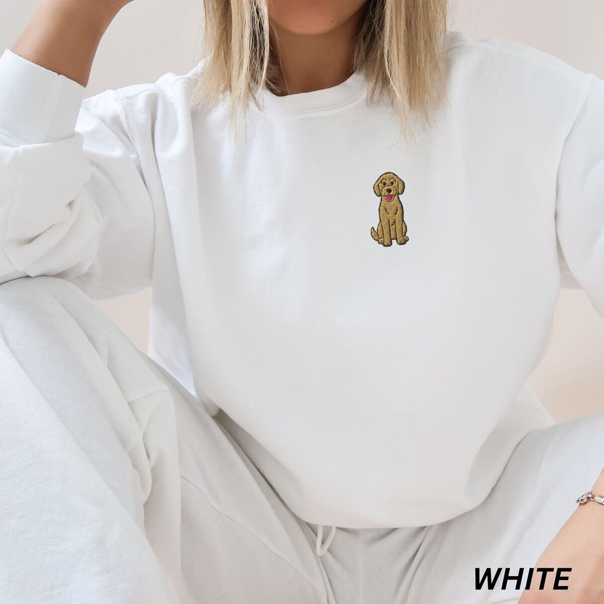 Sitting Goldendoodle Sweatshirt – White Woman in a White crewneck sweatshirt and white joggers, embroidered with a small golden goldendoodle sitting on the left chest