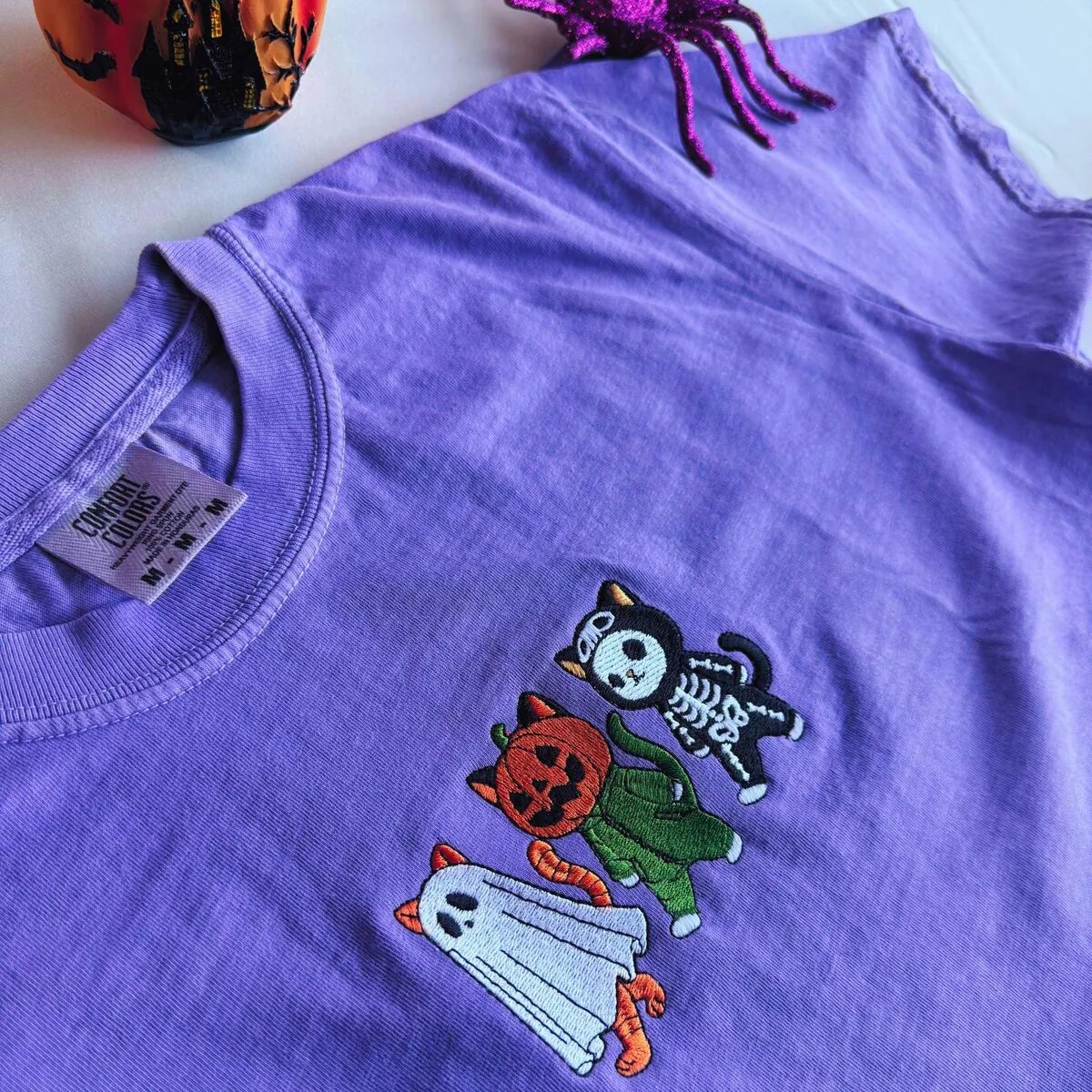 Skeleton Cat, Pumpkin Cat and Ghost Cat Shirt Side view of the embroidered skeleton cat, pumpkin cat and ghost cat on a violet tee