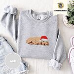 Ash crewneck sweatshirt folded with denim shorts, white sneakers and a potted plant, embroidered with a golden retriever puppy asleep in a red Santa hat, its head resting on its front paws