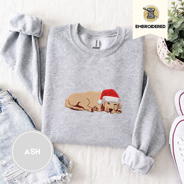 Ash crewneck sweatshirt folded with denim shorts, white sneakers and a potted plant, embroidered with a golden retriever puppy asleep in a red Santa hat, its head resting on its front paws