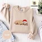 Sand crewneck sweatshirt folded with denim shorts, white sneakers and a potted plant, embroidered with a golden retriever puppy asleep in a red Santa hat, its head resting on its front paws