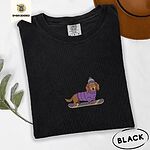 Black Comfort Colors T-shirt embroidered with a long-haired dachshund in a purple sweater and pom-pom beanie riding a snowboard