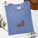 Blue Jean Comfort Colors T-shirt embroidered with a long-haired dachshund in a purple sweater and pom-pom beanie riding a snowboard