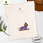 Ivory Comfort Colors T-shirt embroidered with a long-haired dachshund in a purple sweater and pom-pom beanie riding a snowboard