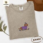 Khaki Comfort Colors T-shirt embroidered with a long-haired dachshund in a purple sweater and pom-pom beanie riding a snowboard