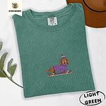 Light Green Comfort Colors T-shirt embroidered with a long-haired dachshund in a purple sweater and pom-pom beanie riding a snowboard