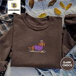 Dark Chocolate crewneck sweatshirt on a plaid flannel shirt with yellow autumn leaves and denim shorts, embroidered with a long-haired dachshund in a purple sweater and pom-pom beanie riding a snowboard