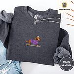 Dark Heather crewneck sweatshirt folded with ripped jeans and a silver chain necklace, embroidered with a long-haired dachshund in a purple sweater and pom-pom beanie riding a snowboard