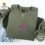Military Green crewneck sweatshirt folded with light jeans and a silver chain necklace, embroidered with a long-haired dachshund in a purple sweater and pom-pom beanie riding a snowboard