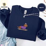 Navy crewneck sweatshirt folded with ripped jeans and a silver chain necklace, embroidered with a long-haired dachshund in a purple sweater and pom-pom beanie riding a snowboard