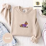 Sand crewneck sweatshirt folded with denim shorts, white sneakers and a potted plant, embroidered with a long-haired dachshund in a purple sweater and pom-pom beanie riding a snowboard