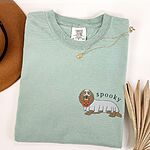 Bay T-shirt laid flat with a necklace and dried palm leaves, embroidered on the left chest with a long-haired dachshund in a ghost sheet and the word spooky