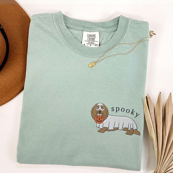 Bay T-shirt laid flat with a necklace and dried palm leaves, embroidered on the left chest with a long-haired dachshund in a ghost sheet and the word spooky