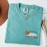 Seafoam T-shirt laid flat with a necklace, embroidered on the left chest with a long-haired dachshund in a ghost sheet carrying a pumpkin bucket under the word spooky