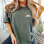 Woman in a sage green T-shirt embroidered on the left chest with a long-haired dachshund in a ghost sheet and the word spooky in white