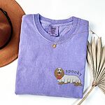 Violet T-shirt laid flat with a necklace and a felt hat, embroidered on the left chest with a long-haired dachshund in a ghost sheet carrying a jack-o'-lantern bucket under the word spooky