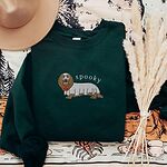 Forest Green crewneck sweatshirt laid flat with a felt hat and dried pampas grass, embroidered in the center with a long-haired dachshund in a ghost sheet and the word spooky in white