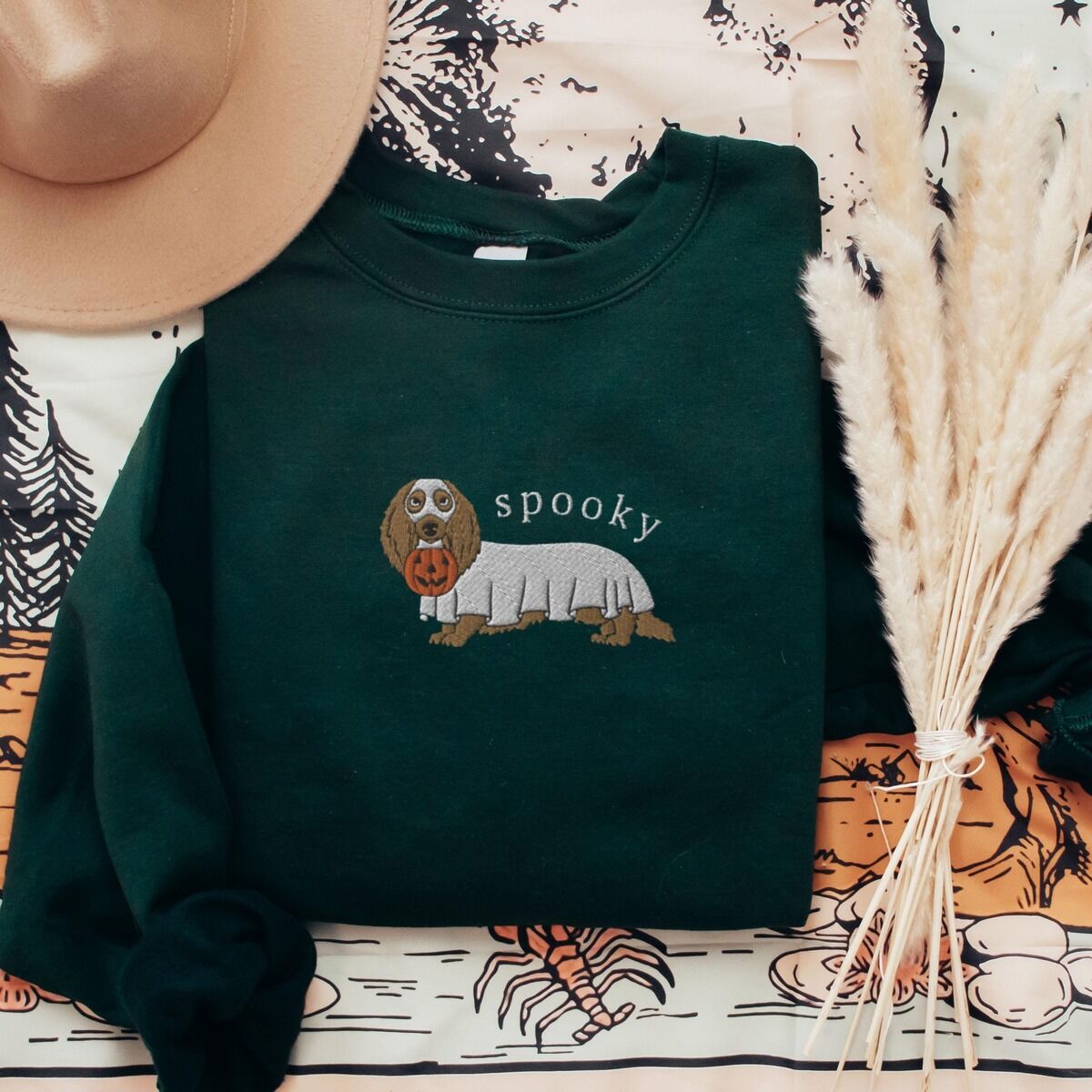 Spooky Dachshund Sweatshirt – Forest Green Forest Green crewneck sweatshirt laid flat with a felt hat and dried pampas grass, embroidered in the center with a long-haired dachshund in a ghost sheet and the word spooky in white