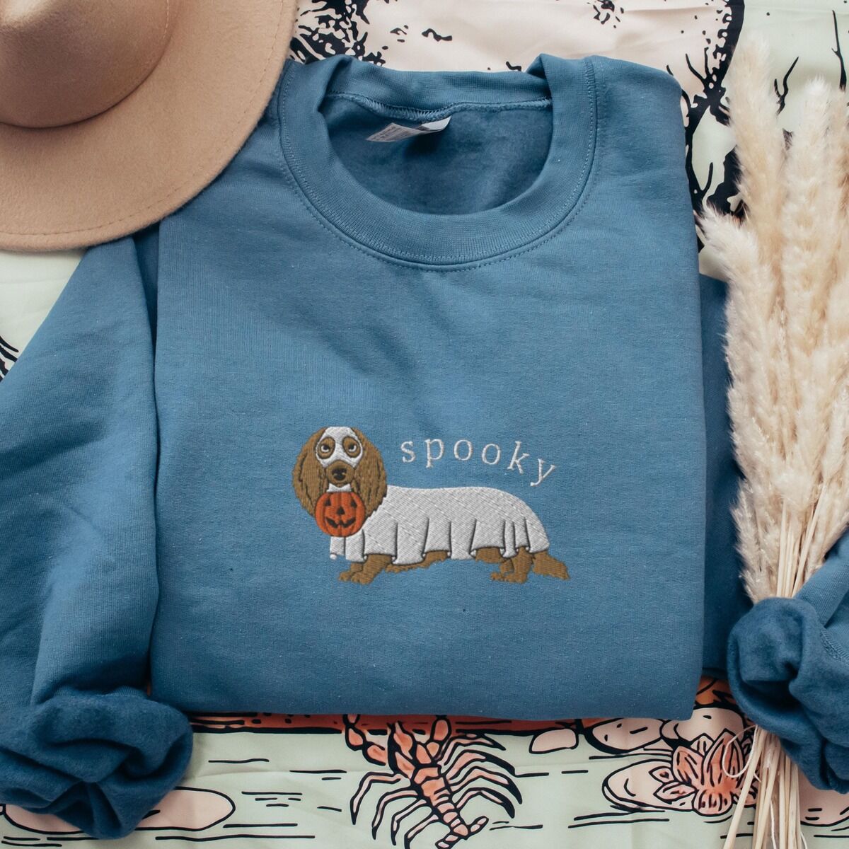 Spooky Dachshund Sweatshirt – Indigo Blue Indigo Blue crewneck sweatshirt laid flat with a felt hat and dried pampas grass, embroidered in the center with a long-haired dachshund in a ghost sheet and the word spooky in white