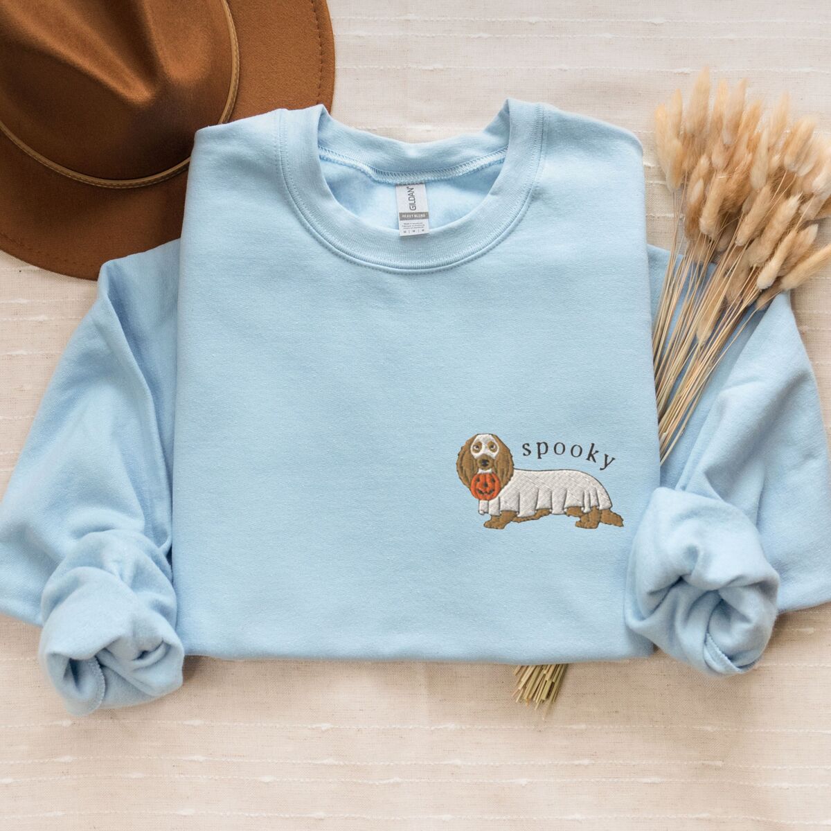 Spooky Dachshund Sweatshirt - Light Blue Light Blue crewneck sweatshirt laid flat with a felt hat and dried wheat, embroidered on the left chest with a long-haired dachshund in a ghost sheet and the word spooky