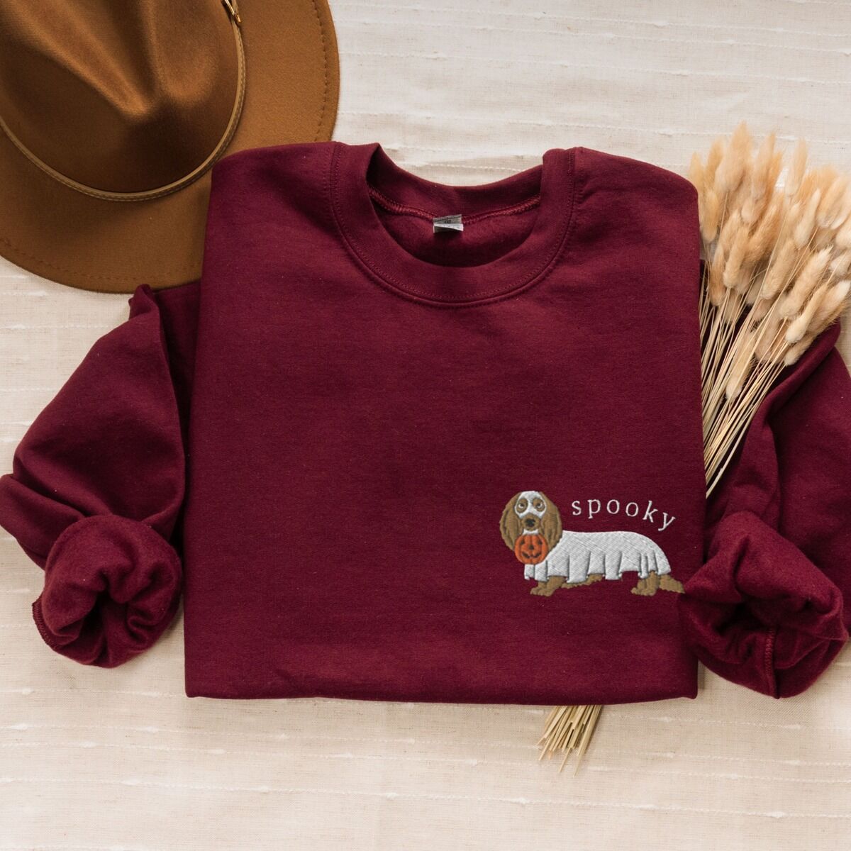 Spooky Dachshund Sweatshirt – Maroon Maroon crewneck sweatshirt laid flat with a felt hat and dried wheat, embroidered on the left chest with a long-haired dachshund in a ghost sheet and the word spooky in white